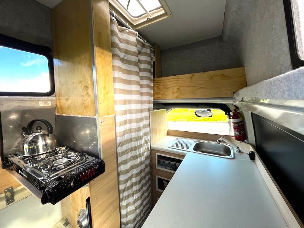 Campervan galley in varnished plywood with a portable two-burner cooker on a fold-down shelf and a striped curtain drawn across the rear