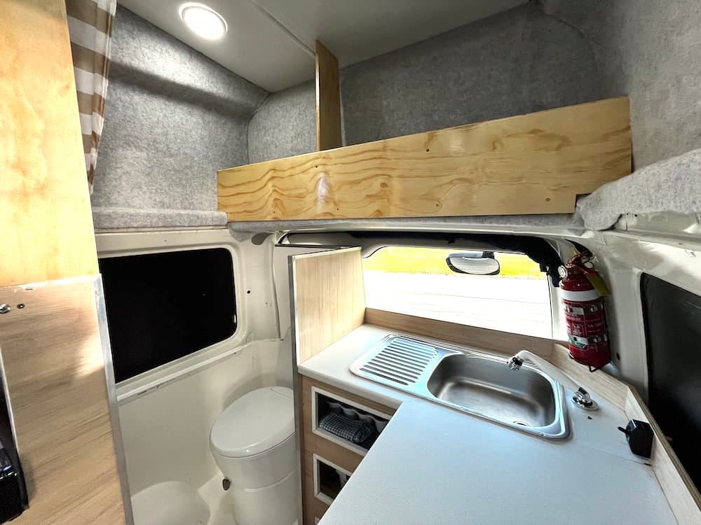 Fixed flush toilet in the corner of the campervan beside a stainless sink and drainer, with open plywood shelving below
