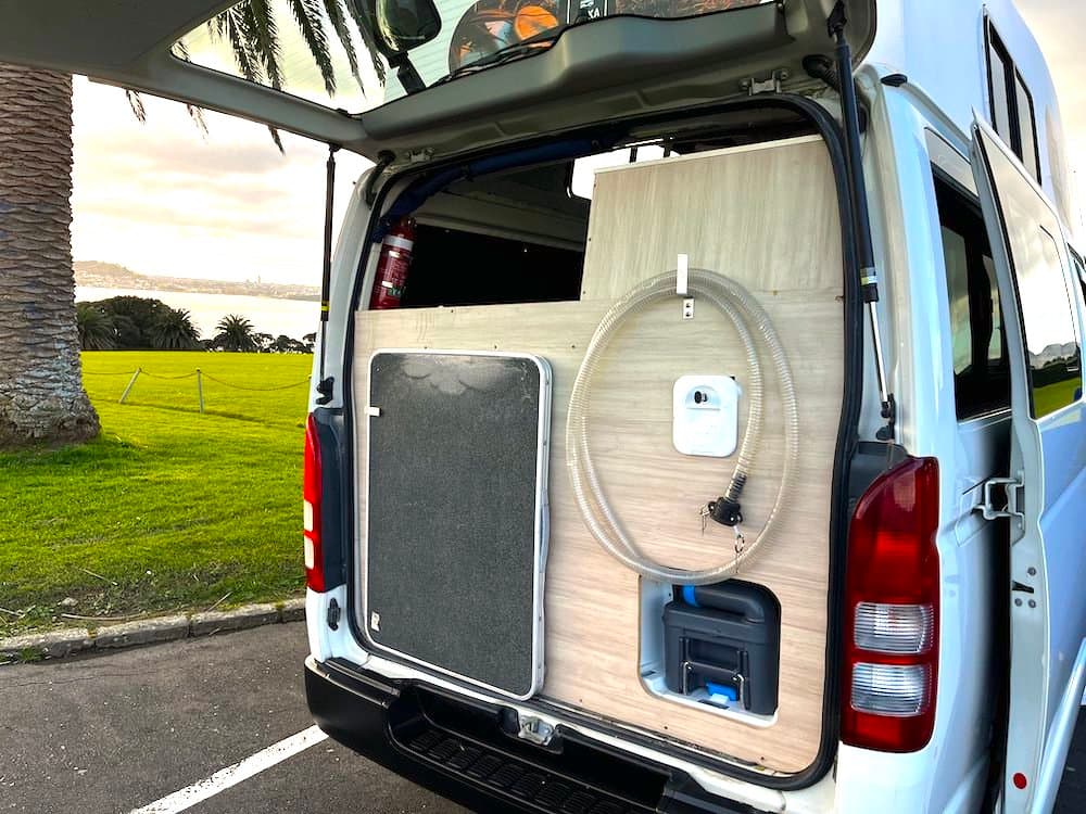 Rear doors of the campervan open, showing a coiled outdoor shower hose and the toilet cassette in its external hatch