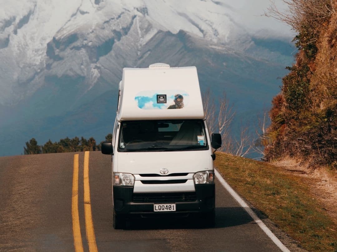 White Toyota HiAce campervan with an over-cab pod driving a sealed road below a snow-covered mountain