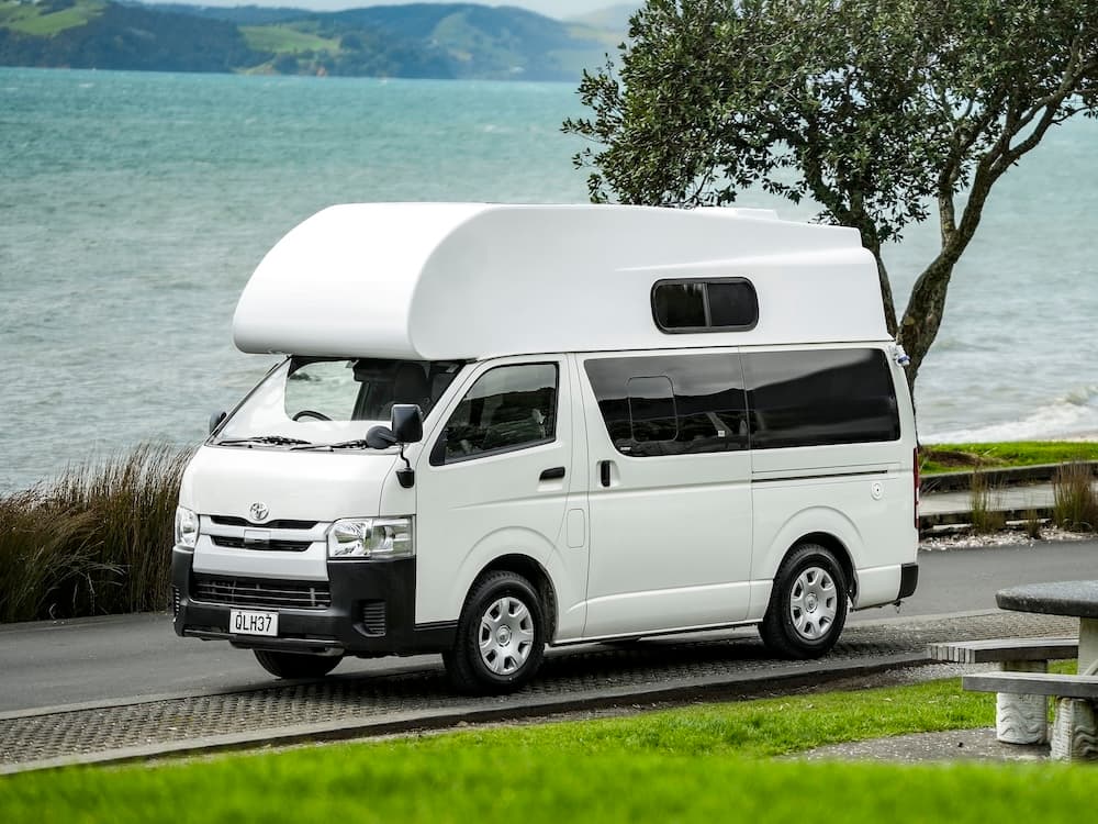 White Toyota HiAce campervan with a fibreglass over-cab pod parked beside the sea