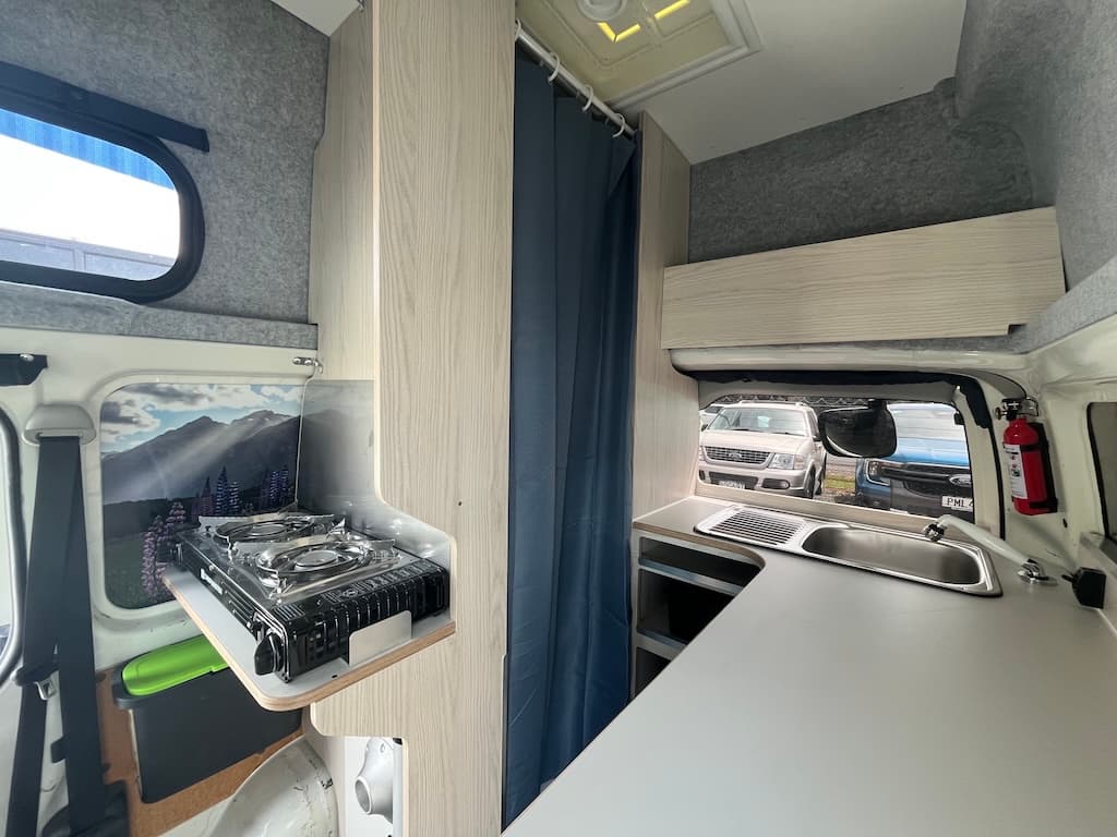 Campervan galley with a portable two-burner gas cooker on a fold-out shelf, a stainless sink and drainer, and a fridge below the bench
