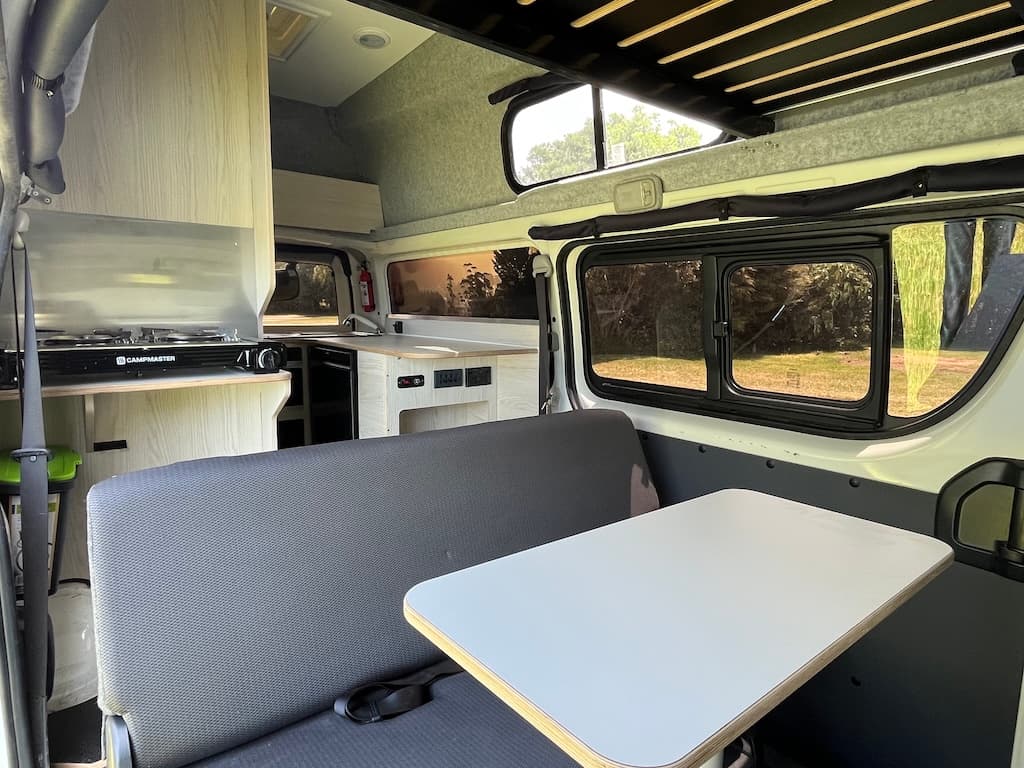 Campervan interior looking forward: upholstered bench, table, two-burner cooker and sink, with the over-cab bed above
