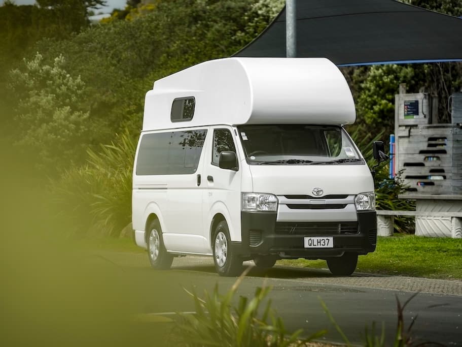 White high-top Toyota HiAce campervan with an over-cab pod, photographed front and side on an open road
