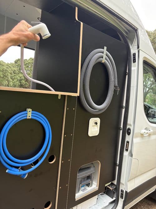 Panel inside the sliding door with a pull-out shower head, a grey waste hose, a blue water hose and a hatch at floor level