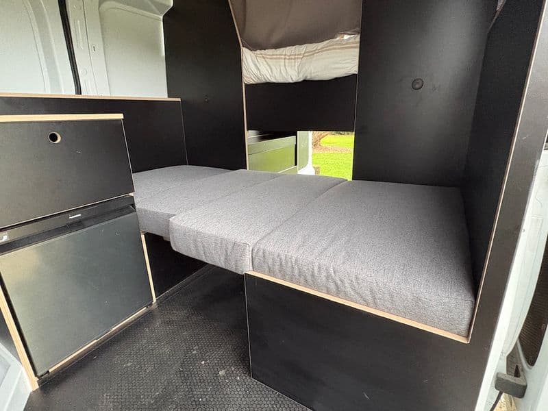 Grey cushions laid across a campervan dinette to form a single bed, with the fixed double bed visible above and behind