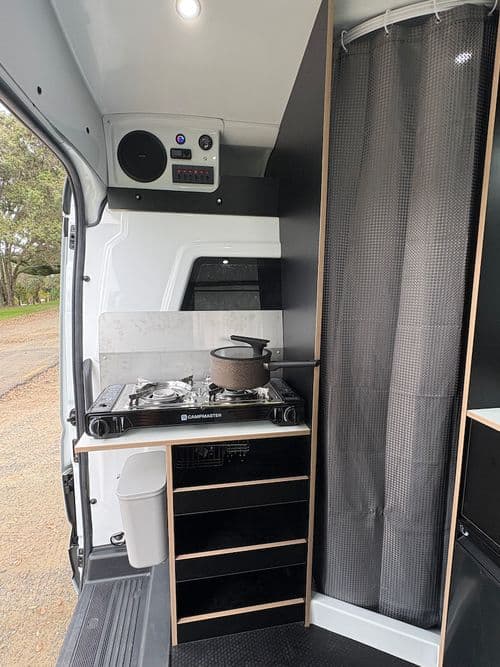 Campervan galley with a portable two-burner gas cooker on a counter, black drawers below, a control panel above and a grey curtain to the right