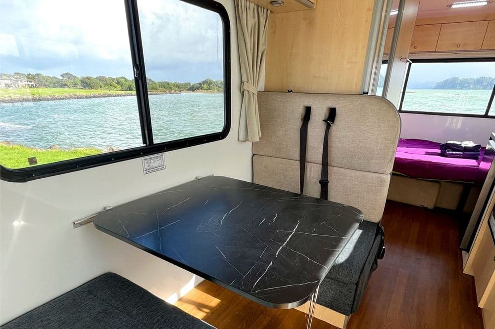 Motorhome living area with a folding dark table, upholstered bench, wood-effect floor and a made bed beyond
