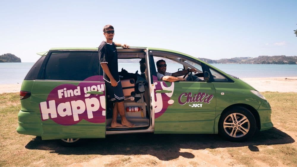 Green Chill'd by JUCY people-mover campervan parked on a beach with its side door open and a man standing in the doorway