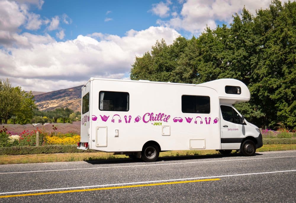 White coachbuilt motorhome in pink Chill'd livery with an over-cab luton, driving a sealed road past a flower field