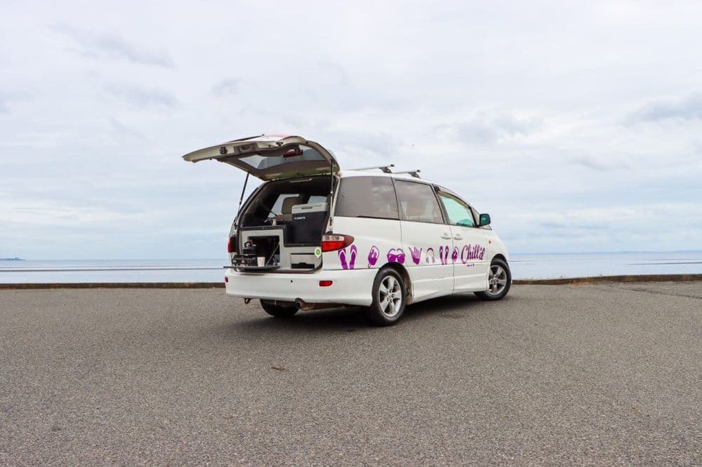 White campervan with its rear hatch raised over a slide-out kitchen unit, parked facing the sea