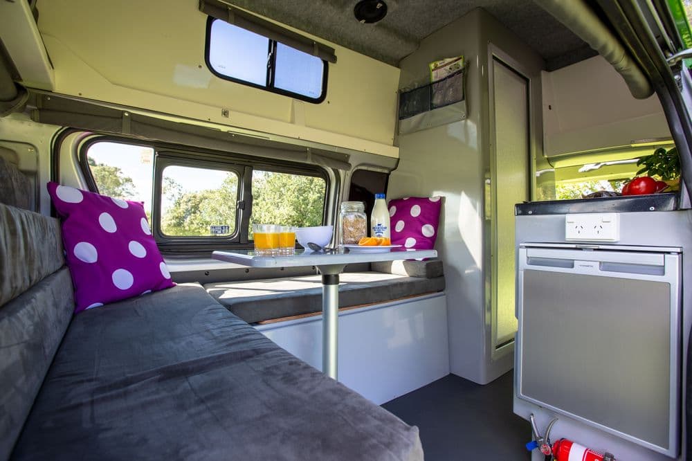 Campervan living area with a grey bench seat, purple polka-dot cushions, a white table, sliding windows and a fridge below the bench