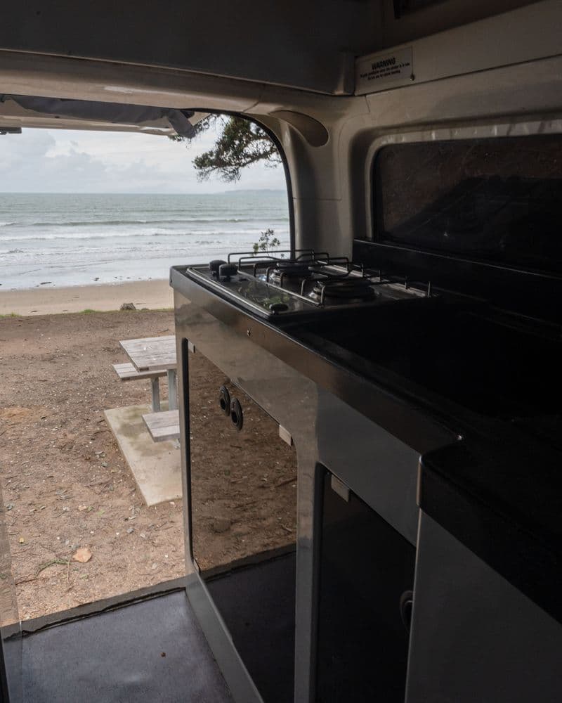 Two-burner gas hob and dark bench in the rear of the campervan, looking out through the open tailgate to a beach