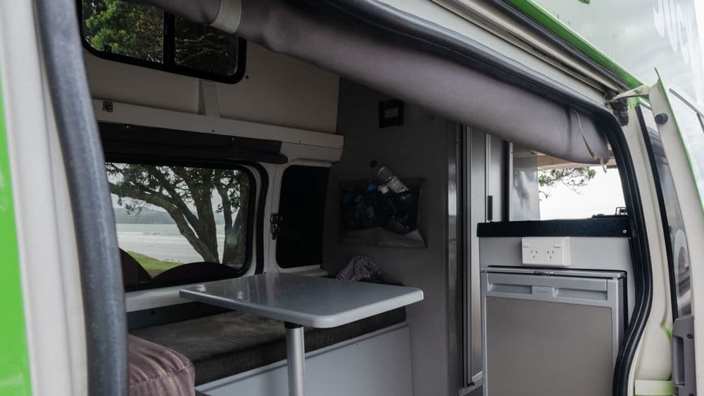 Campervan interior with a bench seat, white table and the fridge below the bench, side door open to trees