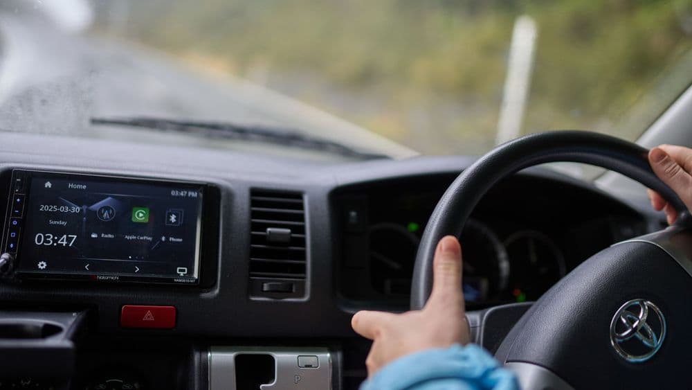Driver's hand on the Toyota steering wheel beside a seven-inch touchscreen showing a media display