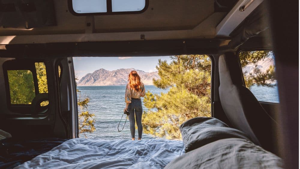 Lower double bed made up in the campervan with the side door open onto a lake shore and a person standing outside