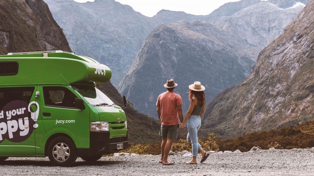 Green campervan with an over-cab pod in JUCY livery parked on gravel at the head of a steep valley, two people walking away from it